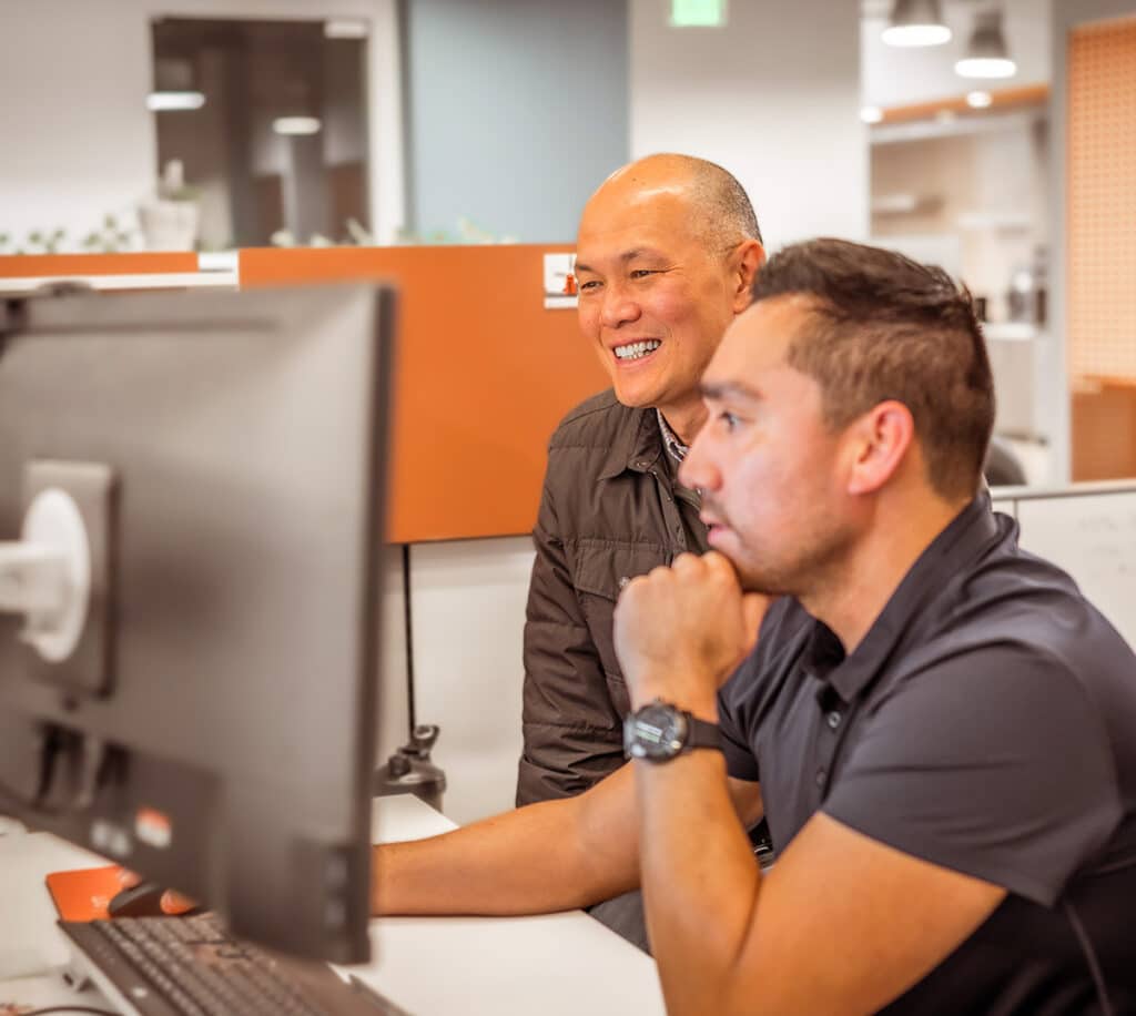 Two men sit at a computer, one is smiling and the other focussing on the computer screen